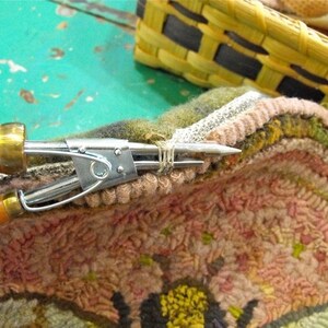 May include: Close-up of a rug hooking tool being used to secure a loop of yarn on a colourful rug. The rug is made of wool and has a variety of colours, including pink, green, and yellow.