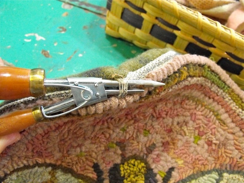 May include: Close-up of a rug hooking tool being used to secure a loop of yarn on a colourful rug. The rug is made of wool and has a variety of colours, including pink, green, and yellow.