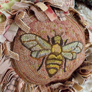 May include: A round, brown and beige rug with a bee design in the centre. The bee is made of yellow, brown and green yarn. The rug is surrounded by a fringe of fabric.