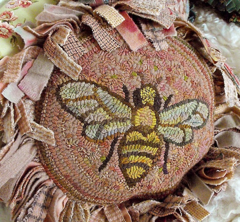 May include: A round, brown and beige rug with a bee design in the centre. The bee is made of yellow, brown and green yarn. The rug is surrounded by a fringe of fabric.
