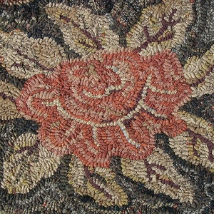 Puede incluir: Una alfombra con un motivo central de rosa creado con una técnica textil de gancho. La rosa está representada en tonos coral y óxido, rodeada de hojas texturizadas en tonos oro, marrón y oliva sobre un fondo oscuro y moteado.
