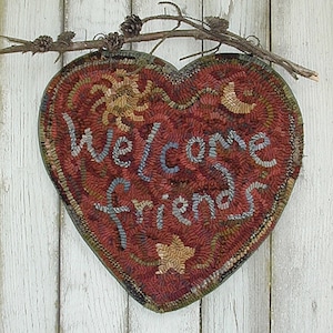 May include: Heart-shaped welcome sign with the words "Welcome Friends" stitched in blue and white. The sign is red with a sun, moon, and star design. A twig with pinecones is attached to the top of the sign. The sign is hanging on a white wooden background.