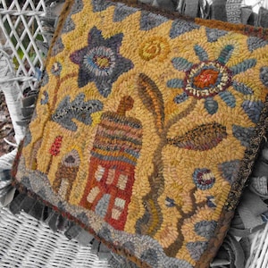 May include: A hand-stitched wool rug pillow with a floral and house design. The pillow features a yellow background with blue, red, and brown accents. The pillow is sitting on a white wicker chair.