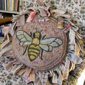 May include: A round, pink and beige rug with a bee design. The rug is surrounded by a fringe of fabric in various shades of beige, pink, and grey.