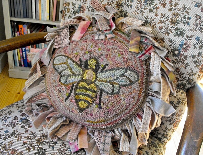 May include: A round, pink and beige rug with a bee design. The rug is surrounded by a fringe of fabric in various shades of beige, pink, and grey.