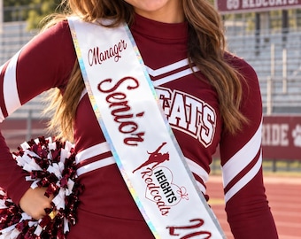 May include: A maroon and white long-sleeved cheerleading uniform with the text CATS on the chest. A white iridescent sash reads Manager Senior Heights Redcoats 2022. A maroon hair bow features the name Linsey and the number 27. Maroon and white pom-poms.