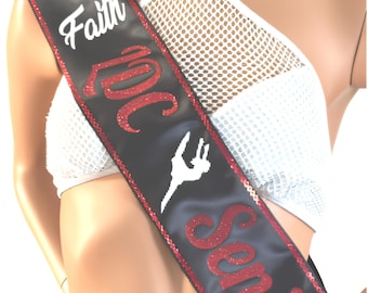 May include: A black satin sash featuring red glitter lettering that reads '2024 Senior' and white script text that reads 'Faith'. A white silhouette of a gymnast performing a leap is centered between the words. The sash has red sequin trim.