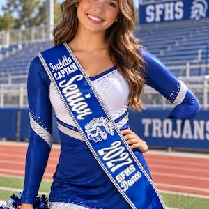 May include: A royal blue and white long-sleeved dance uniform with silver accents and a matching sash. The sash features the text Isabella CAPTAIN Senior 2027 SFHS Dance and a Trojan helmet graphic. The uniform includes a white bodice with rhinestones.