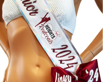May include: A white sash with the words "Manager Senior Heights Redcoats 2024" in maroon lettering. A maroon bow with the name "Linsey" and the number "24" is attached to the sash. The sash is worn over a white bikini top and bottom.