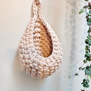 May include: A beige, teardrop-shaped, macrame hanging basket. The basket is made of thick, woven yarn and has a loop at the top for hanging. The basket is empty and hangs against a white wall. A small wooden bead secures the loop.