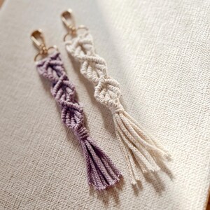 May include: Two macrame keychains. One is a light purple color, and the other is off-white. Both have a woven design with a tassel at the bottom and a gold-colored clasp at the top. The keychains are on a textured, off-white surface.