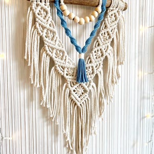May include: A macrame wall hanging with a blue tassel and wooden beads. The macrame is made with natural cotton cord and has a geometric pattern.