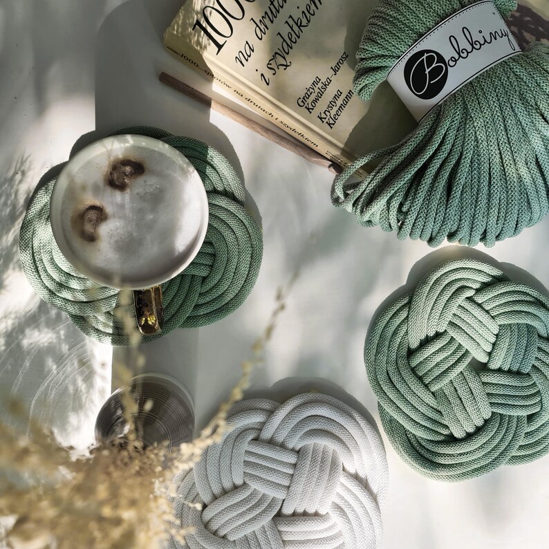 May include: Three green and white crocheted coasters with a ball of green yarn in the background. The yarn ball has a label that says "Bobbiny".