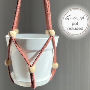 May include: A terracotta-colored macrame plant hanger with wooden beads, holding a white 6-inch pot. "6-inch pot included" is written in the image.