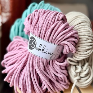 May include: Close-up of several spools of Bobbiny Premium 5mm cotton cord in various colors. The main spool is a soft pink, with others in teal and cream. The label on the pink spool reads "Bobbiny". The cords are tightly wound, suggesting they are for crafting.