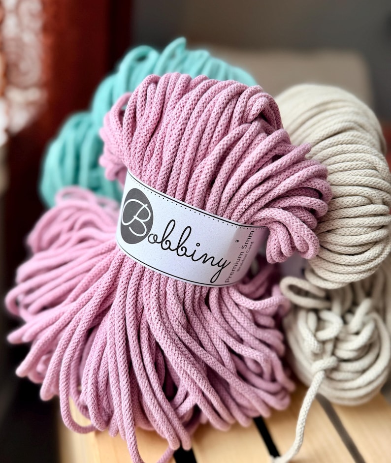 May include: Close-up of several spools of Bobbiny Premium 5mm cotton cord in various colors. The main spool is a soft pink, with others in teal and cream. The label on the pink spool reads "Bobbiny". The cords are tightly wound, suggesting they are for crafting.