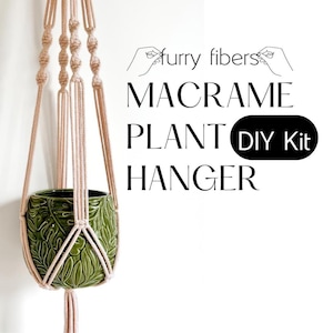 Peut inclure: Un kit de suspension de plante en macramé avec le texte "furry fibers MACRAME PLANT DIY Kit HANGER". Le support est en corde beige et contient un pot en céramique vert avec un motif de feuilles.
