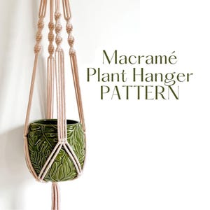 以下が含まれることがあります： ベージュのマクラメプラントハンガーが、葉模様の緑色のセラミックポットを支えています。「Macramé Plant Hanger PATTERN」の文字が濃い緑色で表示されています。プラントハンガーは白い背景に吊り下げられています。