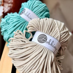 May include: Two skeins of Bobbiny brand macrame cord in teal and beige. Each skein is wrapped with a white label featuring the brand name. The cords are thick and tightly wound, ideal for various crafting projects.