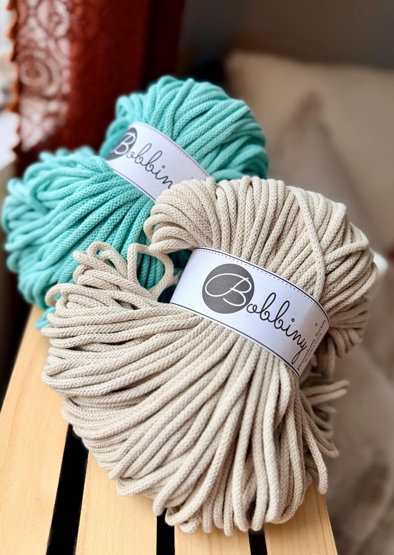 May include: Two skeins of Bobbiny brand macrame cord in teal and beige. Each skein is wrapped with a white label featuring the brand name. The cords are thick and tightly wound, ideal for various crafting projects.