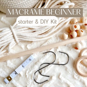 Pode incluir: Um kit de iniciação e DIY para iniciantes em macramê com o texto "MACRAME BEGINNER starter & DIY Kit". O kit inclui corda branca suja, contas de madeira, um pino de madeira, ganchos de metal, uma fita métrica e anéis de madeira. Os itens estão dispostos em uma superfície branca e texturizada.