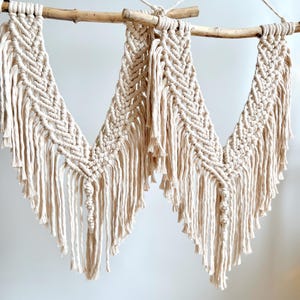 May include: Two macrame wall hangings in a natural cream color, suspended from wooden branches. The woven design features a chevron pattern and long fringe, creating a bohemian aesthetic. The wall decor is hung with twine.