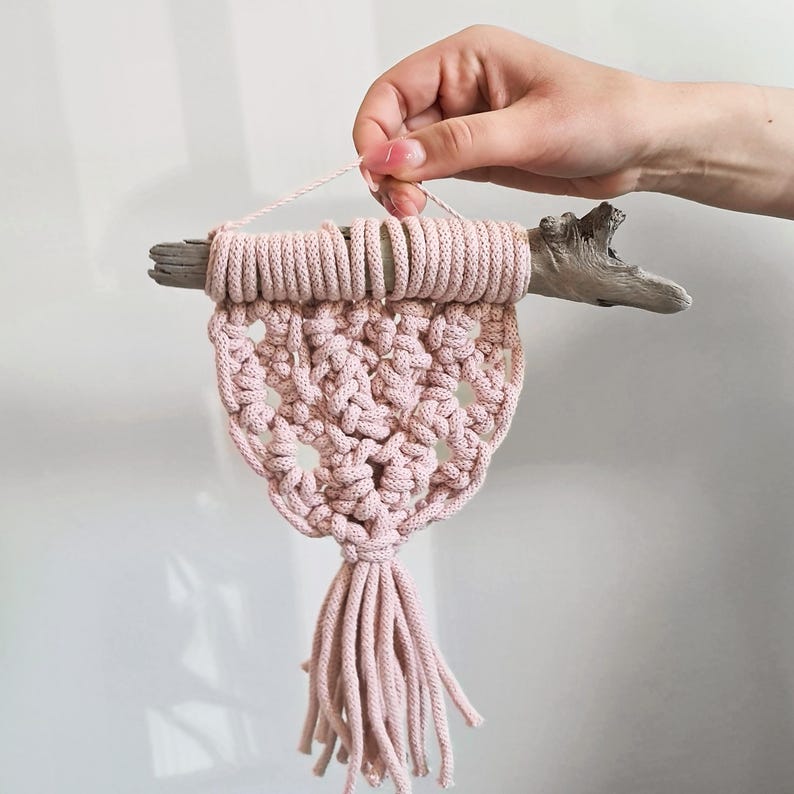 May include: A handmade macrame wall hanging in a soft pink hue. The design features a knotted pattern with a central openwork section and a tassel fringe. It is suspended from a piece of driftwood, adding a natural touch to the decor.