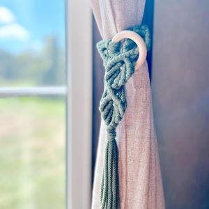 May include: A sage green macrame curtain tieback with a wooden ring and a tassel. The tieback is wrapped around a beige curtain, with a window and a blurred outdoor scene in the background. The macrame design adds a bohemian touch to the window treatment.