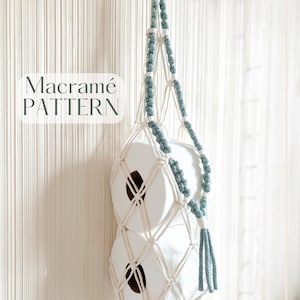 May include: A white macramé toilet roll holder with teal detailing, holding two rolls of toilet paper. The holder is suspended from a hook. The image includes the text "Macramé PATTERN".