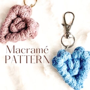 以下が含まれることがあります： 柔らかい白い背景に、2つのマクラメのハート型キーホルダーが表示されています。1つはダスティーローズ、もう1つはライトブルーです。キーホルダーは金属製のリングと留め金に取り付けられています。「Macramé PATTERN」の文字が見えます。