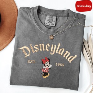 A gray t-shirt with the text "Disneyland EST. 1955" and an embroidered image of Minnie Mouse, a character from the Disney theme park.