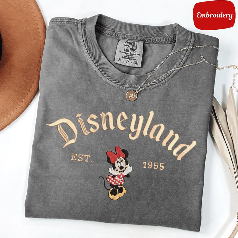 A gray t-shirt with the text "Disneyland EST. 1955" and an embroidered image of Minnie Mouse, a character from the Disney theme park.