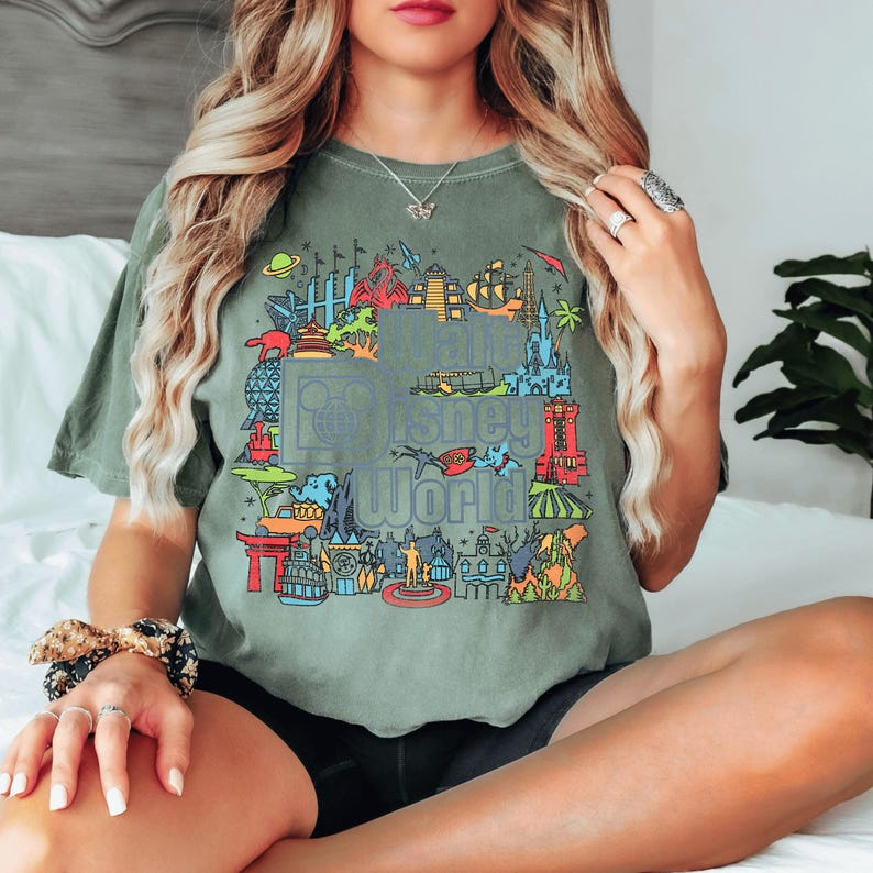A woman with long, wavy blonde hair is sitting on a bed, wearing a green t-shirt with a colorful graphic design.
