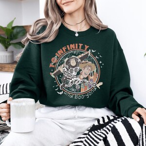 A woman wearing a green sweatshirt with a graphic of characters from the Toy Story franchise, holding a mug and sitting on a striped blanket.