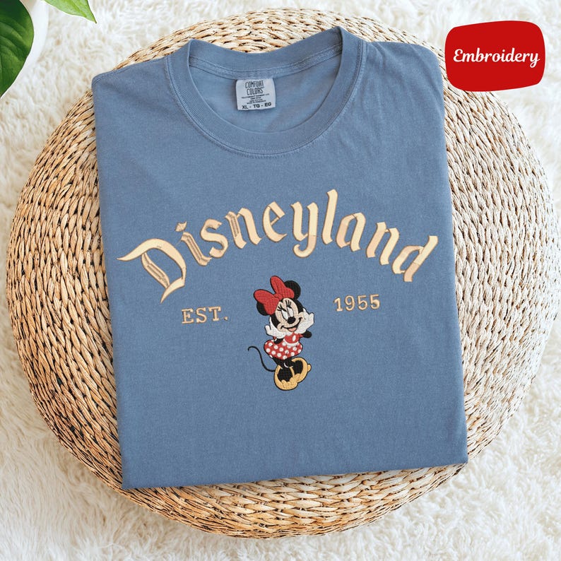 A blue t-shirt with the text "Disneyland EST. 1955" and an embroidered image of Minnie Mouse, a character from the Disney theme park.
