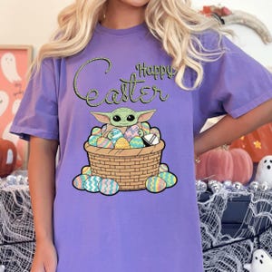 A person wearing a purple t-shirt with a graphic of a young child, likely representing the character Baby Yoda from the Star Wars franchise, sitting in a basket filled with Easter eggs.