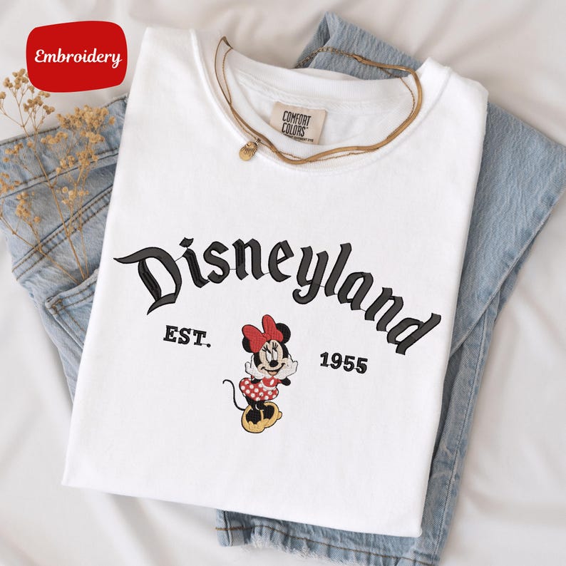 A white t-shirt with the text "Disneyland EST. 1955" printed on it, along with a Minnie Mouse graphic. The t-shirt is placed on top of a pair of blue jeans.
