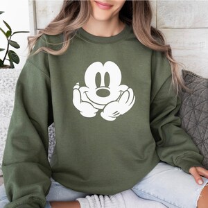 A woman wearing a green sweatshirt with a white Mickey Mouse logo is sitting on a couch.