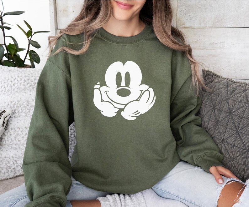 A woman wearing a green sweatshirt with a white Mickey Mouse logo is sitting on a couch.