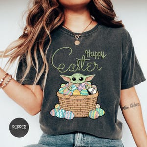 A woman wearing a gray t-shirt with a graphic design of a young child, likely representing the character Baby Yoda from the Star Wars franchise, sitting in a basket filled with Easter eggs.