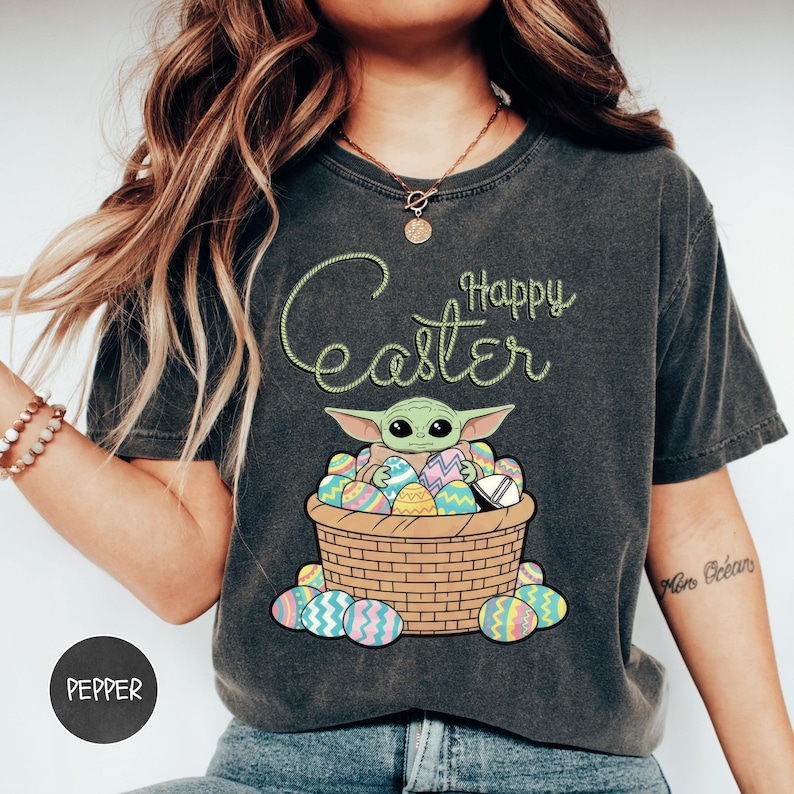 A woman wearing a gray t-shirt with a graphic design of a young child, likely representing the character Baby Yoda from the Star Wars franchise, sitting in a basket filled with Easter eggs.