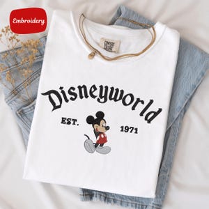A white t-shirt with the text "Disneyworld EST. 1971" printed on it, along with a Mickey Mouse graphic. The t-shirt is placed on top of a pair of blue jeans.