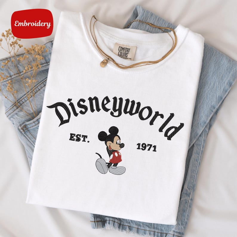 A white t-shirt with the text "Disneyworld EST. 1971" printed on it, along with a Mickey Mouse graphic. The t-shirt is placed on top of a pair of blue jeans.
