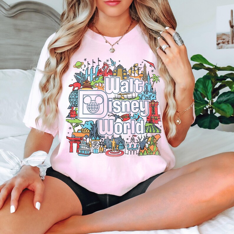A woman wearing a pink t-shirt with a graphic of Disney World landmarks and the text "Walt Disney World" is sitting on a bed.