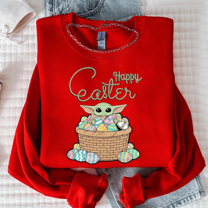 A red sweater with a graphic of a young child, likely representing the character Baby Yoda from the Star Wars franchise, sitting in a basket filled with Easter eggs. The sweater has the text "Happy Easter" printed on it.
