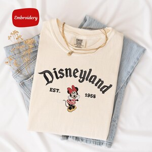 A white t-shirt with the text "Disneyland EST. 1955" printed on it, along with a Minnie Mouse graphic, is laid out on a white surface.