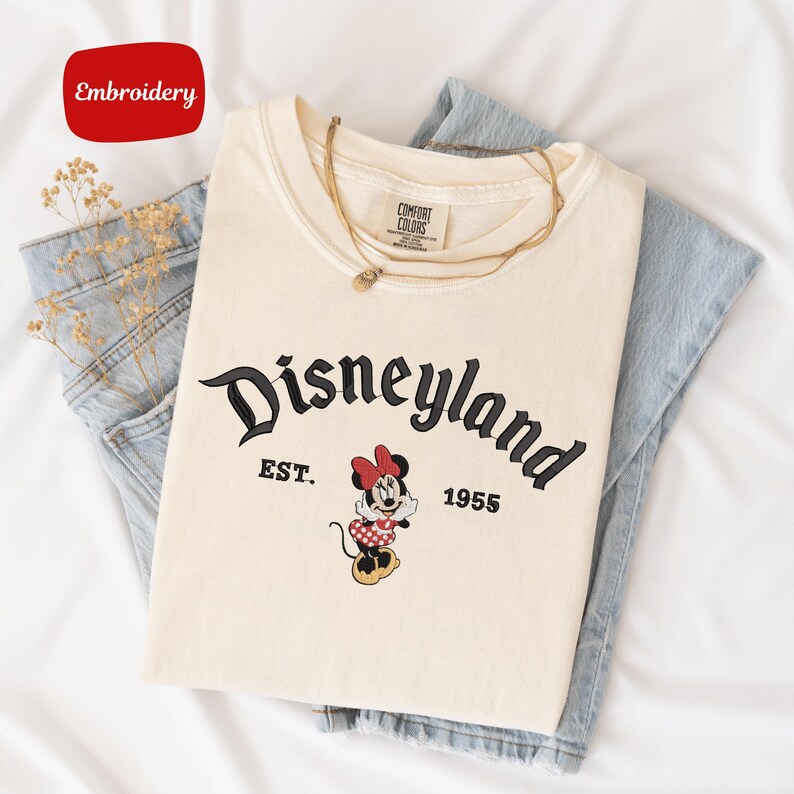 A white t-shirt with the text "Disneyland EST. 1955" printed on it, along with a Minnie Mouse graphic, is laid out on a white surface.
