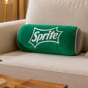 A green throw pillow with the word "Sprite" printed on it is placed on a beige couch, with a wooden coffee table in the foreground and a plant in a glass vase on the table.