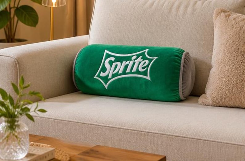 A green throw pillow with the word "Sprite" printed on it is placed on a beige couch, with a wooden coffee table in the foreground and a plant in a glass vase on the table.