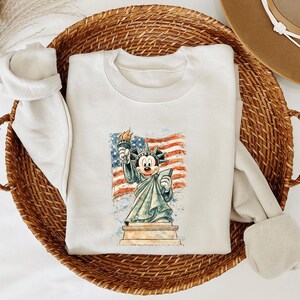A white sweatshirt with a cartoon image of Mickey Mouse holding the American flag is displayed in a woven basket.